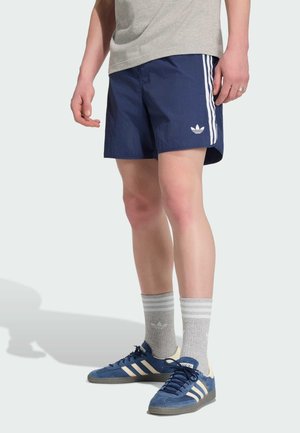 Person trägt marineblaue Adidas-Shorts mit weißen Streifen, graue Adidas-Socken und blaue Adidas-Sneaker und steht vor hellem Hintergrund.