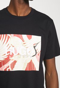 Sort t-shirt med en rød, hvid og lyserød rektangulær grafik af en flyvende trane og "HUGO" tekst på brystet.