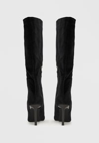Fekete térdig érő magassarkú csizma, amely bársonyból készült, sima textúrával, karcsú tűsarkúval és oldalsó cipzár részlettel.