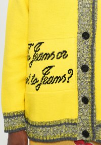 Gul stickad cardigan med en svart textdesign, grå mönstrad kant och svarta knappar. Har en framficka med broderad detalj.