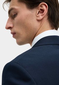 Marineblauw colbert met een gestructureerde schouder, witte overhemdkraag, gladde stof en strakke lijnen, van opzij gepositioneerd.