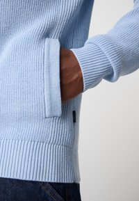 Light blue knitted sweater with ribbed cuffs and hem. Features a side pocket and woven label. Soft texture, relaxed fit, and attention to detail.
