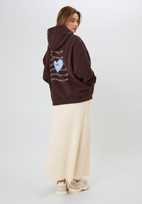 Sudadera grande marrón con un gráfico de un corazón azul en la parte de atrás, combinada con una falda larga color crema y zapatillas beige.