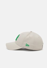 Gorra beige con visera curva, que presenta un logo bordado verde y acentos. Tejido de algodón con orificios para ventilación y transpirabilidad.