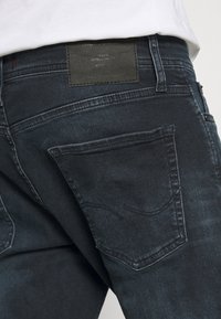 Jeans azul escuro com um tecido texturizado, design de cinco bolsos, costura em contraste e um patch de couro na parte de trás da cintura.