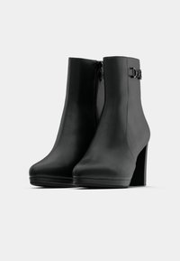 Botas de couro pretas até ao tornozelo, com bico arredondado, salto bloco alto, fecho de correr lateral e um detalhe decorativo de fivela na parte externa do tornozelo.