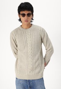 Suéter de punto en beige claro con cuello redondo, patrones texturizados y puños y dobladillo acanalados, combinado con jeans de mezclilla azul.