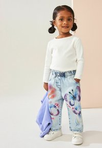 Jeans din denim de culoare bleu deschis cu motive florale în nuanțe de roz și violet, asociați cu o cămașă albă cu mânecă lungă și adidași albi.