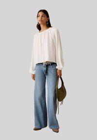 Weiße Bluse mit langem Arm, gerafftem Ausschnitt und seitlichen Knöpfen, kombiniert mit hellblauen Schlagjeans und einer grünen Schultertasche.