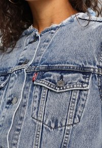 Ljusblå denimjacka med fransad krage, silverfärgade knappar, två bröstfickor och ett litet rött logomärke på den vänstra fickan.