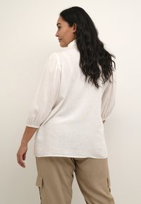 Blouse en lin blanc avec manches bouffantes, coupe décontractée et texture subtile. Assortie à un pantalon cargo kaki avec poches latérales.