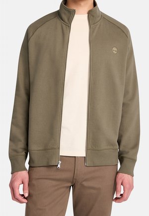 Homme portant une veste zippée vert olive sur une chemise beige et un pantalon marron, debout avec les mains détendues sur les côtés.