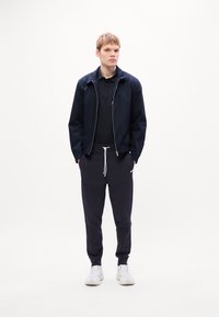 Ung mand iført en marineblå jakke, sort polo, sorte joggers med hvide snore og hvide sneakers, der står foran en simpel hvid baggrund.