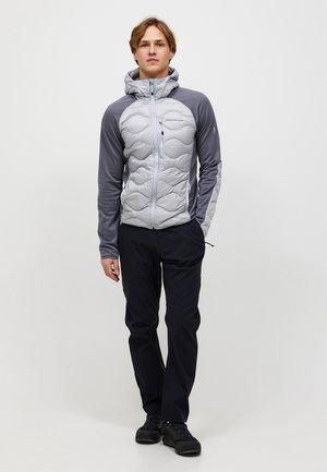 Młody mężczyzna stoi, ubrany w jasno szarą pikowaną kurtkę z ciemnoszarymi rękawami i czarne spodnie na jednolitym białym tle.