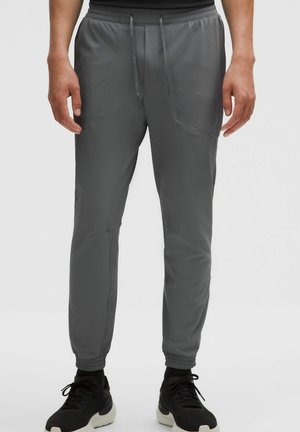 Osoba nosząca szare, teksturowane spodnie dresowe ze ściągaczami przy nogawkach i ściągaczem w pasie, w połączeniu z czarno-białymi trampkami.