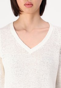 Sweter dziany w kolorze białym z dekoltem w kształcie V, o luźnym, teksturowanym splotem. Posiada żeberkowy dekolt i długie rękawy, prezentując otwarty splot.