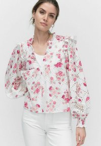 Hvid bluse med blomsterdesign i pink roser, flæser på skuldrene og blonder detaljer på de pufede ærmer. Letvægts, semi-gennemsigtig stof.