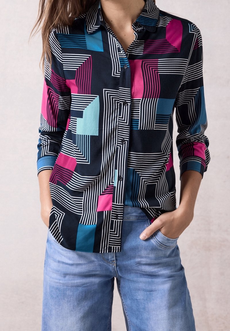Kobieta w koszuli na guziki z długim rękawem z geometrycznymi wzorami w kolorach czarnym, różowym, niebieskim i białym, w połączeniu z jasnoniebieskimi dżinsami.