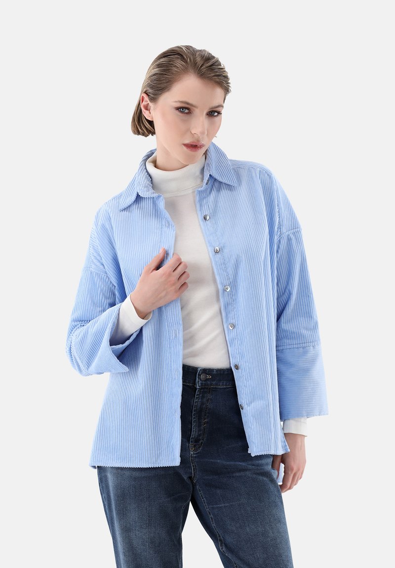 Blaue Cordhemdjacke mit Knopfleiste, Umlegekragen und lockerer Passform. Getragen über einem weißen Rollkragenpullover und dunklen Jeans.