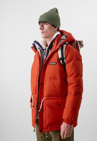 Oransje pufferjakke med hette, med fleece-fôret krage, frontlommer og snor. Brukt med en olivengrønn lue og ryggsekk.