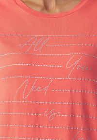 Koraal t-shirt van katoenmix stof, met zilveren strasslettering en horizontale lijnen, wat een textuurdetail creëert.