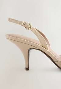 Beige korkokenkä, jossa on ohut nilkkahihna ja kultainen solki, sileä, pyöreä korko vaalealla taustalla.