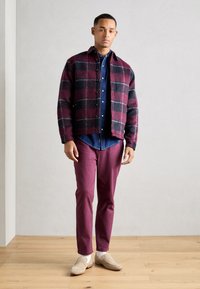 Veste à carreaux bordeaux et noir, chemise bleue en dessous, et pantalon bordeaux. Des mocassins beiges complètent la tenue. Fond en bois.