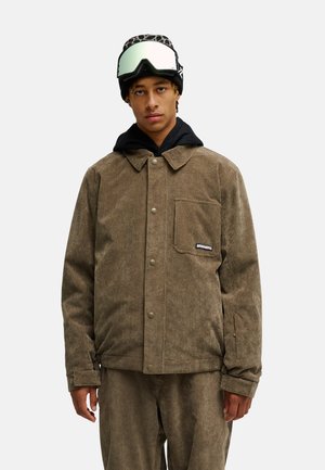 Junger Mann trägt eine braune Cordjacke und Hose, schwarzen Hoodie, Leopardenmütze und Skibrille auf der Stirn, steht vor weißem Hintergrund.