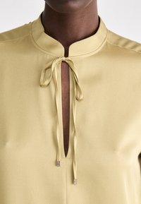 Blusa gialla chiara con colletto alzato, dotata di scollatura a chiave e chiusura a fiocco. Tessuto liscio e dettagli di design minimalisti.