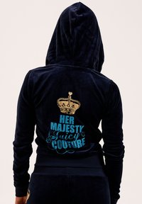 Veste de survêtement en velours marine avec capuche, ornée d'un graphique de couronne dorée et du texte turquoises "SA MAJESTÉ Juicy COUTURE" au dos.