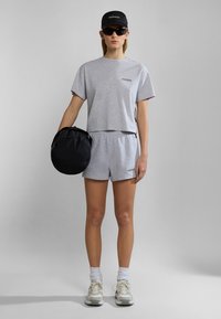 Grijze cropped t-shirt en shorts, van katoen, gecombineerd met een zwarte pet en zonnebril. Zwarte tas in de hand; witte sneakers met grijze accenten.