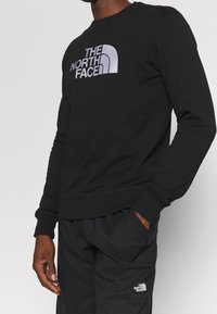 Svart bomullströja med en silverfärgad logotyp på bröstet, ribbade ärmslut och nederkant. Kombineras med svarta byxor som ger en avslappnad passform.