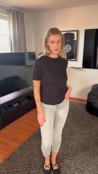 Mladá žena s blond vlasmi, čiernym tričkom, svetlými džínsami a čiernymi topánkami stojaca v obývacej izbe s televízorom a zarámovanými umeleckými dielami.