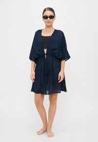 Kimonou transparent de culoare albastru marin, cu mâneci largi, talie cu șnur și croială lejeră. Asortat cu un top negru fără mâneci și purtat cu picioarele goale.