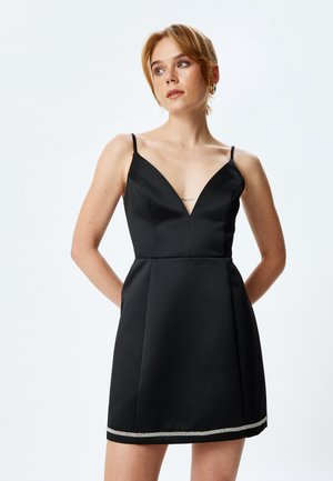 Schwarzes ärmelloses Kleid mit tiefem V-Ausschnitt, figurbetontem Oberteil und schwingendem Rock. Verfügt über einen mit Kristallen verzierten Saum für zusätzliche Details.