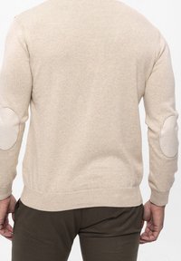 Beige trui met een gestructureerde brei. Heeft contrasterende elleboogpatches en een ribbelzoom. Mouwen zijn aansluitend met een ronde manchette.