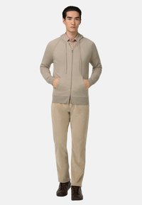 Beige mikina na zip s kapucí na stahování a předními kapsami, spárovaná se světle béžovými kalhotami a tmavými botami, vytváří casual outfit.