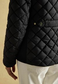 Čierna prešívaná bunda s diamantovým vzorom, s detailom gombíka na chrbte a textúrovaným, vypchávkovým povrchom. Pod bundou vidieť biele nohavice.