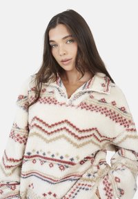 Polarowy pulower z wysokim kołnierzem, z ciepłą kremową podstawą, w zygzakowate wzory w kolorze czerwonym, beżowym i niebieskim oraz z suwakiem na pół. 