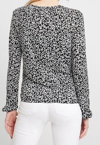 Blusa con motivo floreale in bianco e nero, con maniche lunghe, dotata di un collo arricciato e polsini frangiati, indossata con jeans bianchi.