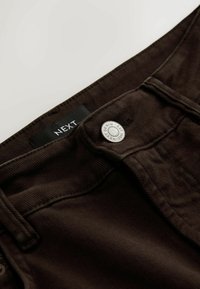 Pantaloni din denim de culoare maro închis, cu o textură ribbed, având un nasture argintiu și o etichetă neagră pe care scrie "NEXT" pe talie.