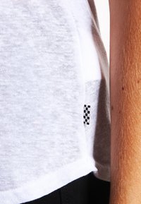 Bílo-šedý pruhovaný tričko, vyrobené z lehkého materiálu, má zakřivený lem a malou černo-bílou šachovnicovou nášivku na boku.