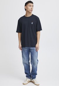 Navy blue cotton t-shirt with short sleeves, featuring a small graphic design on the chest. Paired with light blue denim jeans and white sneakers.