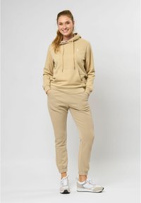 Beige hettegenser med lomme foran og snor, kombinert med matchende avsmalnende joggebukser og lyse joggesko.