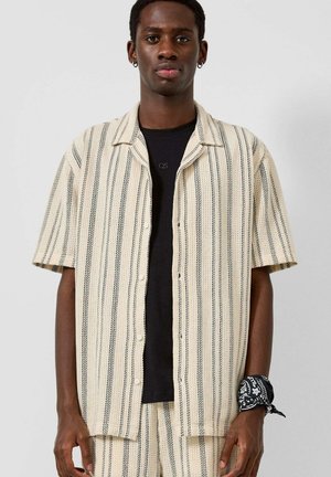 Młody mężczyzna w beżowej i czarnej koszuli w pionowe paski z krótkim rękawem, na czarnej koszulce, z dopasowanymi spodniami w paski i czarną bandaną na nadgarstku.