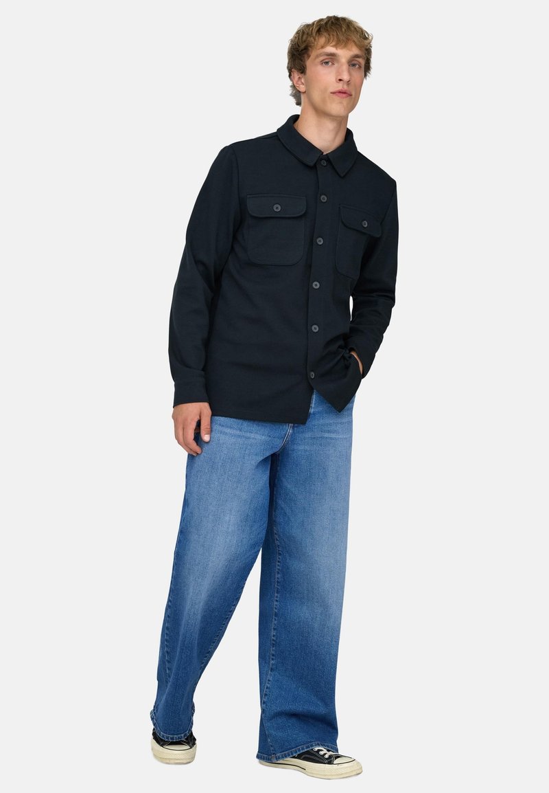 Junger Mann, der ein schwarzes Hemd mit Brusttaschen, weit geschnittene blaue Jeans und schwarze Turnschuhe mit weißen Sohlen trägt.