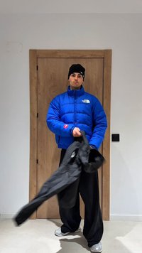 Blaue Steppjacke mit hohem Kragen, mit einem Logo auf der Brust, kombiniert mit weiten schwarzen Hosen und grauen Turnschuhen.