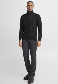 Fekete cipzáras nyakú pulóver, texturált kötött anyagból készült, sötétszürke, slim-fit nadrággal és csattal ellátott fekete cipővel párosítva.