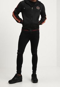 Muž v černé mikině na zip s červenými a béžovými pruhy na rukávech, černých otrhaných úzkých džínách a černých teniskách, stojící s rukama v kapsách.