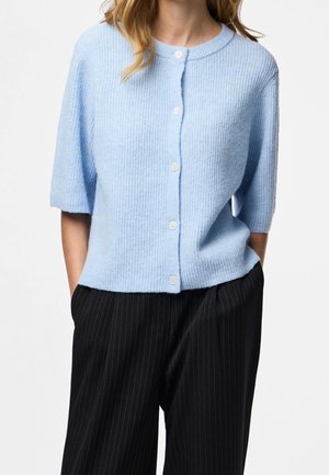 Kvinde iført en lyseblå kortærmet strikket cardigan med knapper og sorte brede bukser med smalle striber, hænder i lommerne.